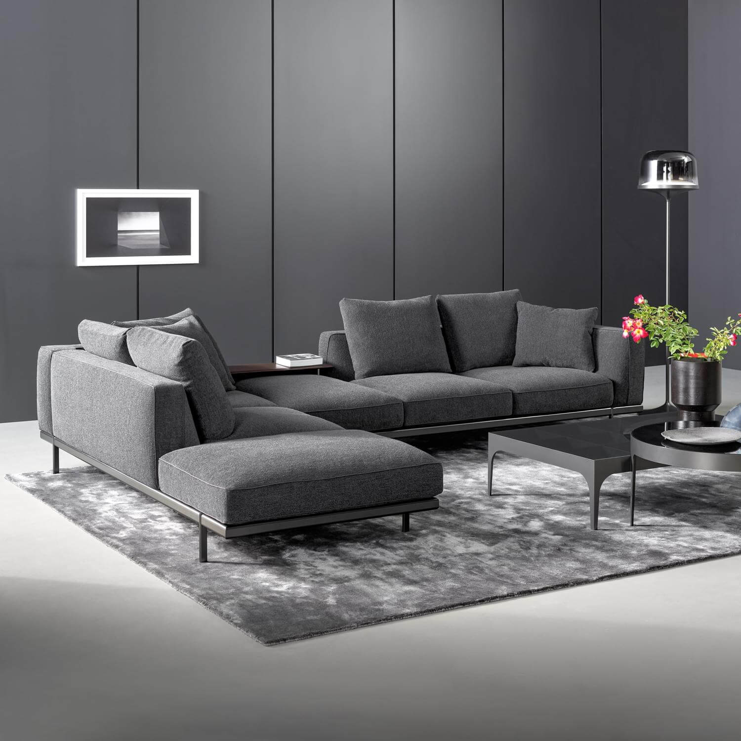 Modular sofa dark gray fabric chaise lounge, metal legs, gray paneled wall, floor lamp, glass coffee table, textured rug, floral arrangement.