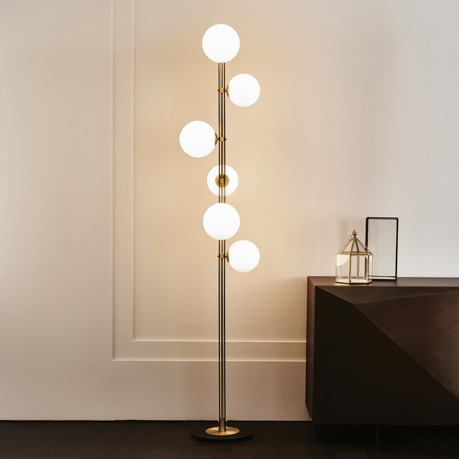 Cattelan Italia Planeta floor lamp, brushed brass finish, six opaque white glass globes suspended from a slender stem against a beige paneled wall.