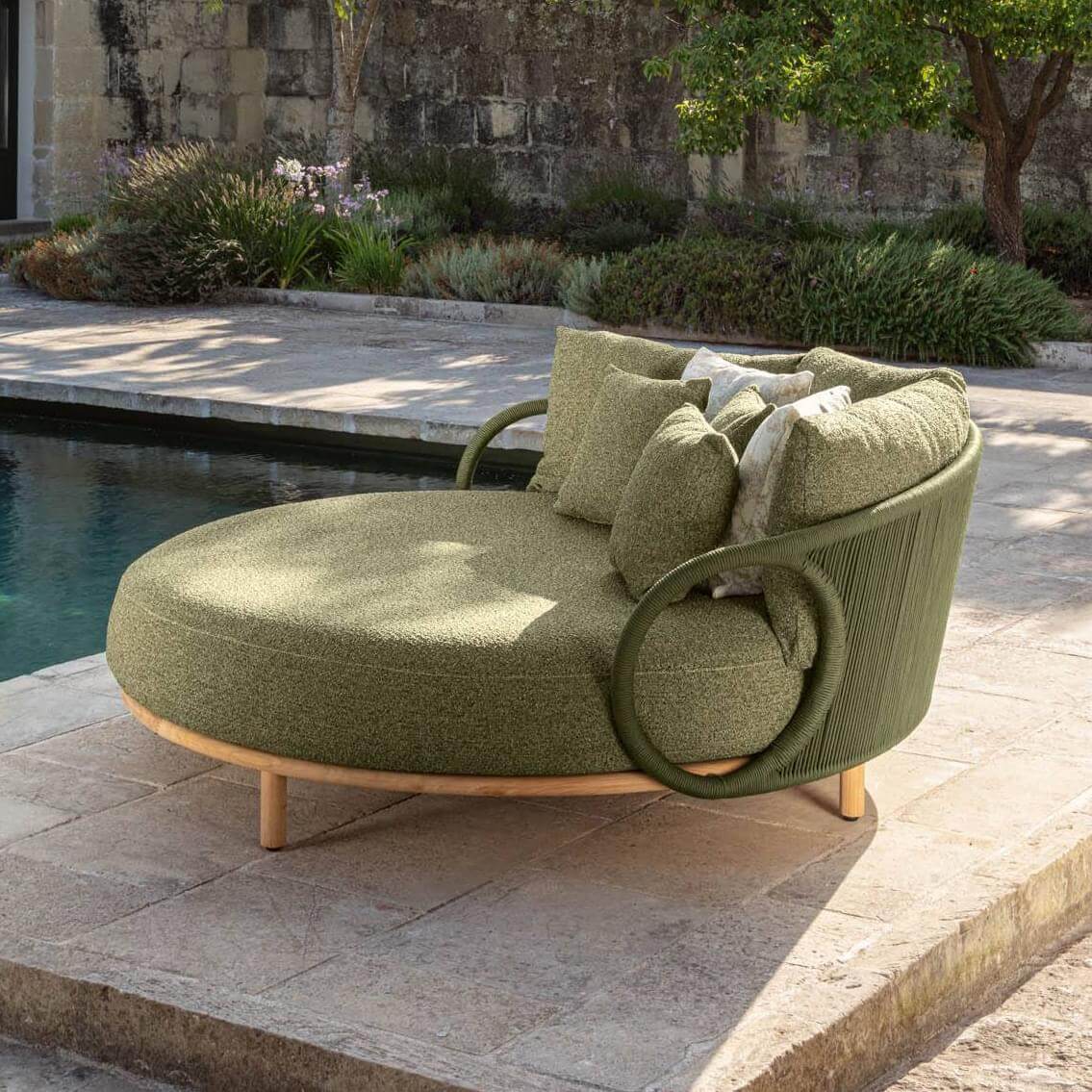 Olive-green circular chaise lounge with rope detail, supported by light-wood legs, and styled with textured pillows on a stone patio.