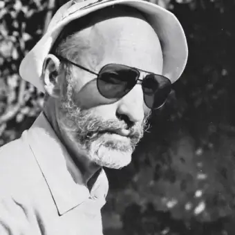 Grayscale portrait of a man with a hat, sunglasses, and full beard facing slightly left against a blurred background