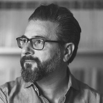Grayscale portrait of a man with dark hair, beard, and glasses looking to the side against a blurred rectangular background