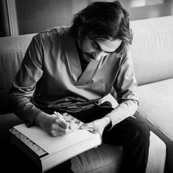 Man with dark hair seated and leaning over an open book with visible photographic pages and a pen