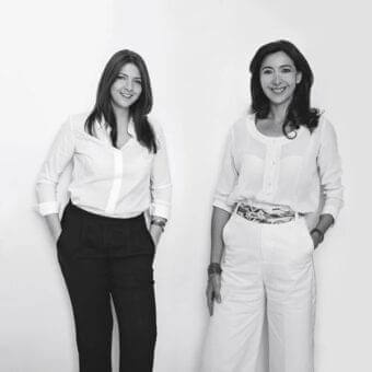 Two women in light shirts and dark trousers stand facing forward with slight smiles and visible bracelets