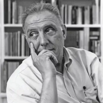 Gray scale portrait of a man with gray hair resting his chin on his hand before a full bookcase