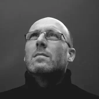 Monochromatic portrait of a man with glasses wearing a dark scarf and jacket against a light gray backdrop