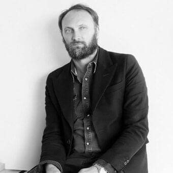 Seated man with beard and dark jacket looks forward against a light gray smooth background