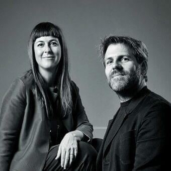 Two people seated close together gaze forward against a plain medium gray background