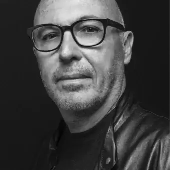 Middle-aged man with shaved head and glasses looks forward wearing a dark t-shirt and leather jacket against a dark background