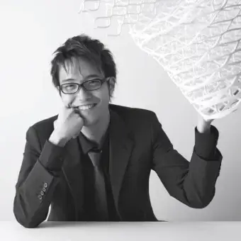 Grayscale portrait of a person in a suit touching their chin beside a large white abstract sculpture with curving shapes