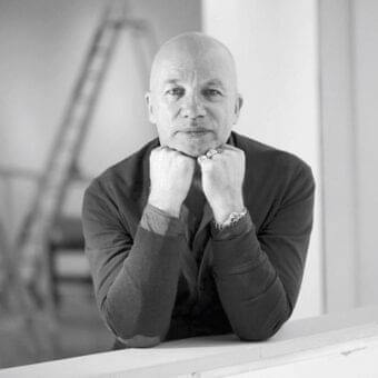 Central grayscale portrait of a man with clasped hands, wearing knitwear and a collared shirt against a blurred architectural background