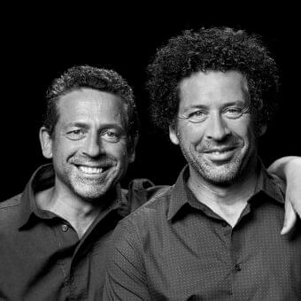 Two men face forward smiling, both wearing collared shirts against a dark grayscale background