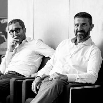 Black armchairs with curved backs sit against a white wall, featuring two men in button-down shirts; partial poster text reads "VERME" and "HAR".