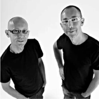 Two men with short hair and glasses wear dark shirts against a pale, neutral backdrop in a symmetrical composition