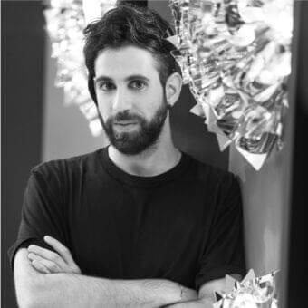 Grayscale portrait of a bearded man with crossed arms, facing forward with blurred reflective shapes in the background