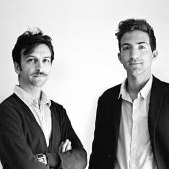 Two people stand facing forward against a plain light gray backdrop with soft, diffused lighting and minimal shadows