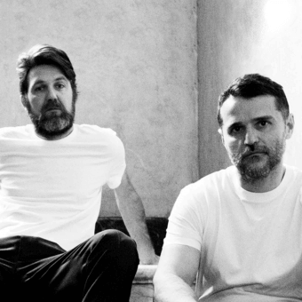 Two men with facial hair sit near a low surface against a light gray mottled wall in monochrome