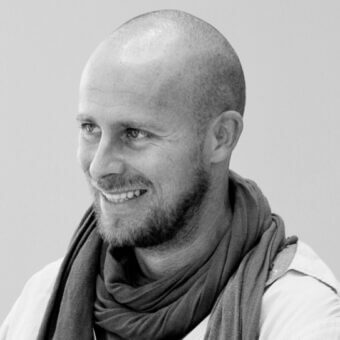 Man portrait, grayscale, shaved head, short beard, brown scarf, light shirt, gentle smile, studio lighting, plain gray background.