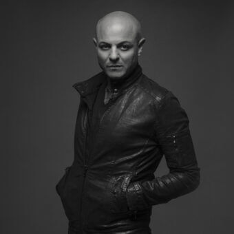 Bald man Alessandro La Spada in black leather jacket, angled pockets, tailored fit, neutral expression, dark gray background.