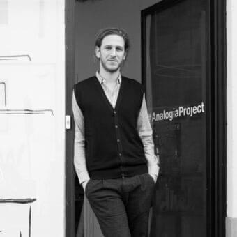 Man in dark cardigan stands near a doorway with glass panel displaying stacked text and architectural markings