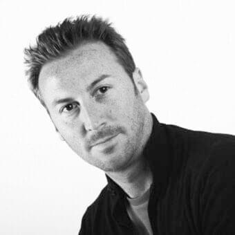 Monochromatic portrait of a man with short dark hair, stubble, and a slight smile, wearing dark and light clothing layers