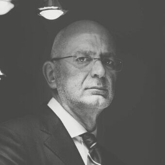 Bald man in dark suit and glasses looks downward with serious expression against dark blurred background