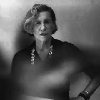 Older person with short hair facing left wearing dark clothing and jewelry against a blurred light background