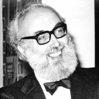 Monochrome portrait of a smiling man with a full white beard and glasses wearing a dark jacket and bow tie against a blurred background
