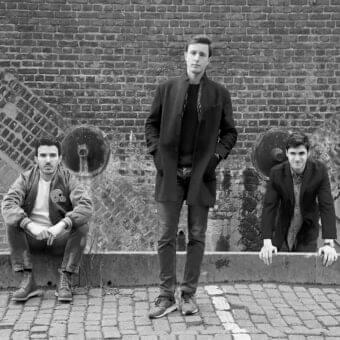 Three people stand and sit against a textured brick wall with circular recesses and a concrete ledge