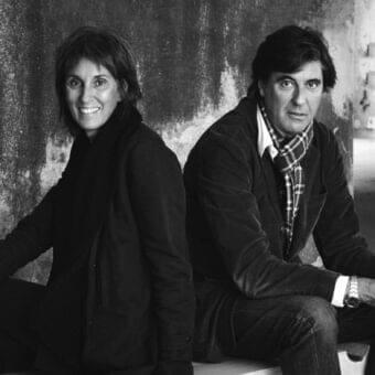 Two seated people face forward against a dark, mottled wall with subtle lighting and soft shadows