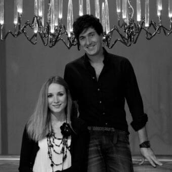 Two people stand near each other beneath a large, curving fixture with upward-pointing elongated shapes against a neutral backdrop