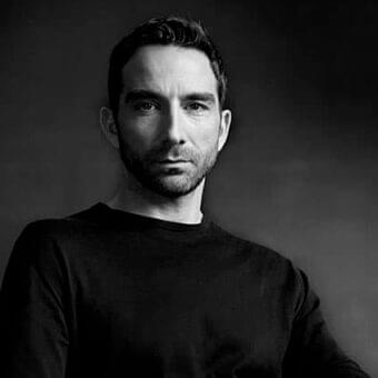 Monochromatic portrait of a man with dark hair and beard looking forward against a dark gray gradient background