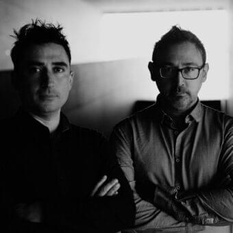Two men with crossed arms look directly ahead against a vertically paneled light-toned wall in grayscale