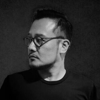 Black and white portrait of Ben Wu wearing glasses and black t-shirt, against a dark gray textured background with dramatic lighting.