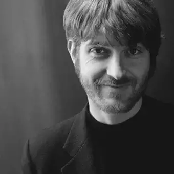 Grayscale portrait of a man with short dark hair and beard, wearing a dark garment against a gradient background