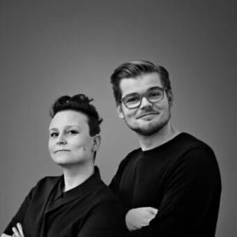 Two people with crossed arms stand close together against a solid gray monochrome background
