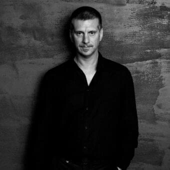 Man with short hair and mustache wearing a dark shirt looks forward against a mottled dark gray textured backdrop