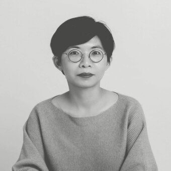 Grayscale portrait of a person with short dark hair and round glasses wearing a textured knitted top against a light background