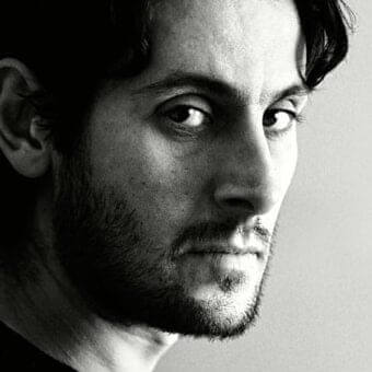 Monochrome portrait of a man facing right with dark wavy hair, beard, and intense gaze against a light gray background