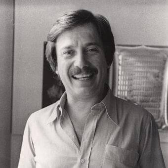 Grayscale portrait of a smiling man with a mustache wearing a light shirt against a softly blurred interior background