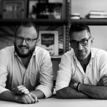Two people with glasses and collared shirts lean on a light surface before a dark shelving unit displaying framed objects and a labeled item