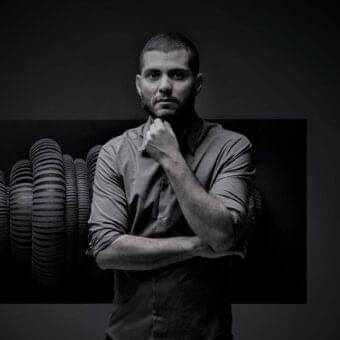 Grayscale portrait of a person with crossed arms against a dark gradient backdrop and stacked cylindrical forms to the left
