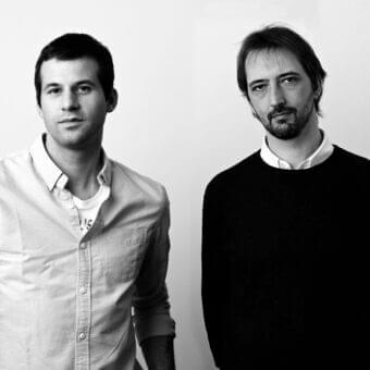Two men facing forward with neutral expressions wear contrasting light and dark clothing against a gray monochromatic backdrop