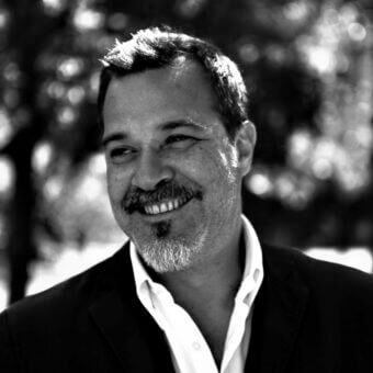 Dario Curatolo portrait, black and white, man in suit jacket and white shirt, mustache, beard, dappled light, blurred foliage background.