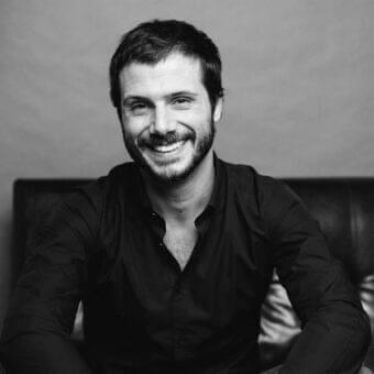 Gray scale portrait of a smiling man with dark hair and beard wearing a dark collared shirt against a softly textured light background