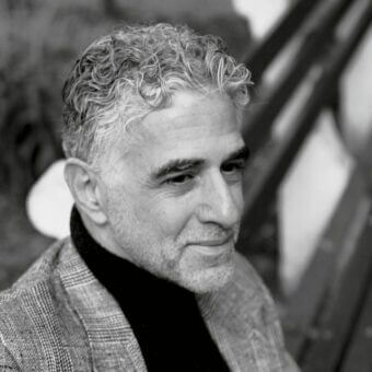 Black and white portrait of David Estreich wearing a black turtleneck and plaid herringbone blazer, with curly hair and a thoughtful expression.