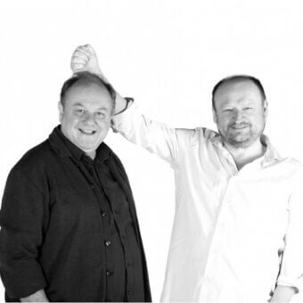 Black and white studio portrait of two smiling men, one in a dark blazer, the other in a white shirt, with a plain backdrop.