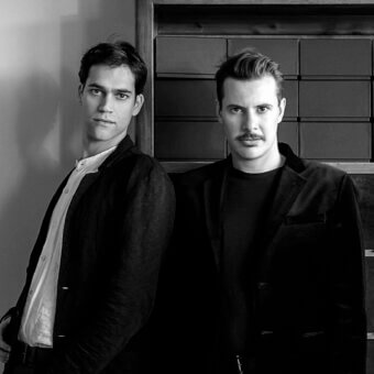 Two men with contrasting hair styles wear dark jackets and light shirts against a grid patterned backdrop