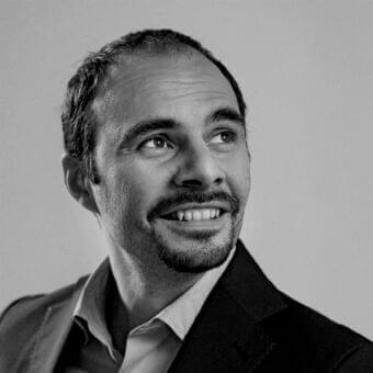 Grayscale portrait of a smiling man with short dark hair, beard, and collared shirt wearing a dark jacket against a light gray background