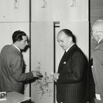 Gallery scene with beige panels displaying sketched figures, a card reading “Mostra di Disegni di Estel,” and three suited men in conversation.