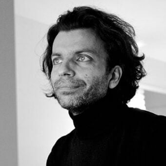 Black and white portrait of a man with wavy dark hair, a trimmed beard, and a black turtleneck, looking off-camera with soft, diffused lighting.